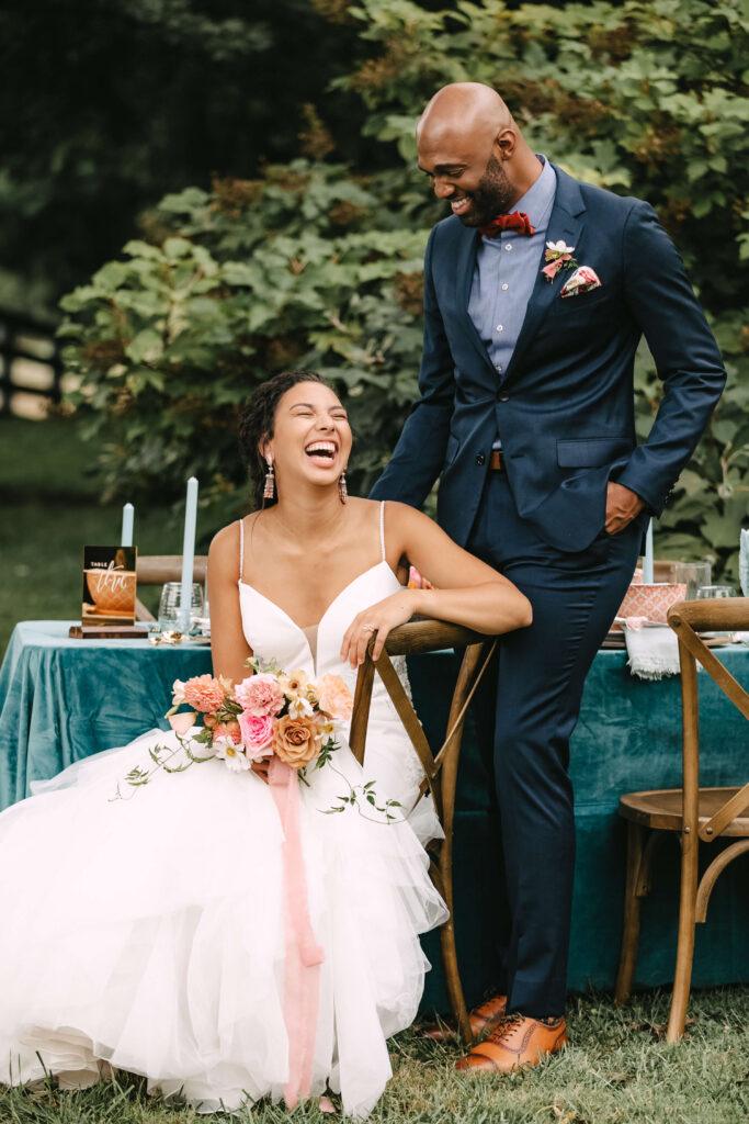 Black couple joyfully celebrates their wedding day outside in front of a beautifully style table in jewel tones. The bride wears a blush pink dress, with a garden bouquet tied with pink ribbon. The groom wears a deep blue suit, with mahogony wingtip shoes. He looks down at her smiling as she sits in a chair laughing up at him. They are incredibly happy.
