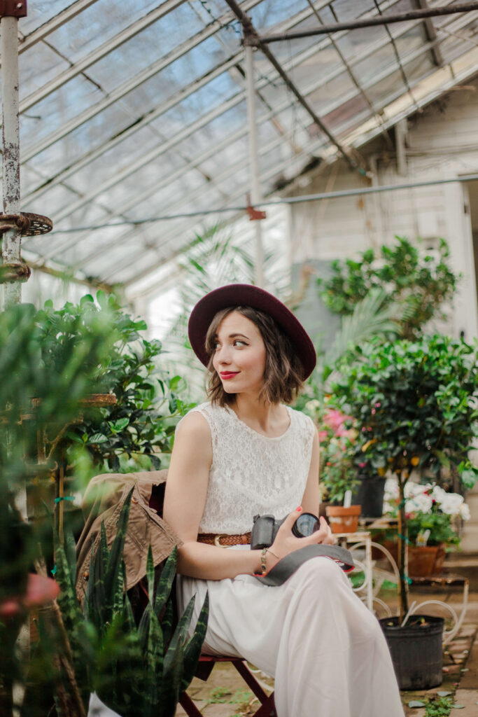 A woman with a secret sits in a green house surrounded by plants. She holds a camera, looking over her left shoulder at something that has caught her eye. A slight grin graces her face, she wears a stylish hat, gently set on the back of her head so that we can clearly see her face.